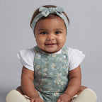 Bebé niña sonriente con overol verde menta estampado floral, camiseta blanca con volantes y diadema de EmpiBy
