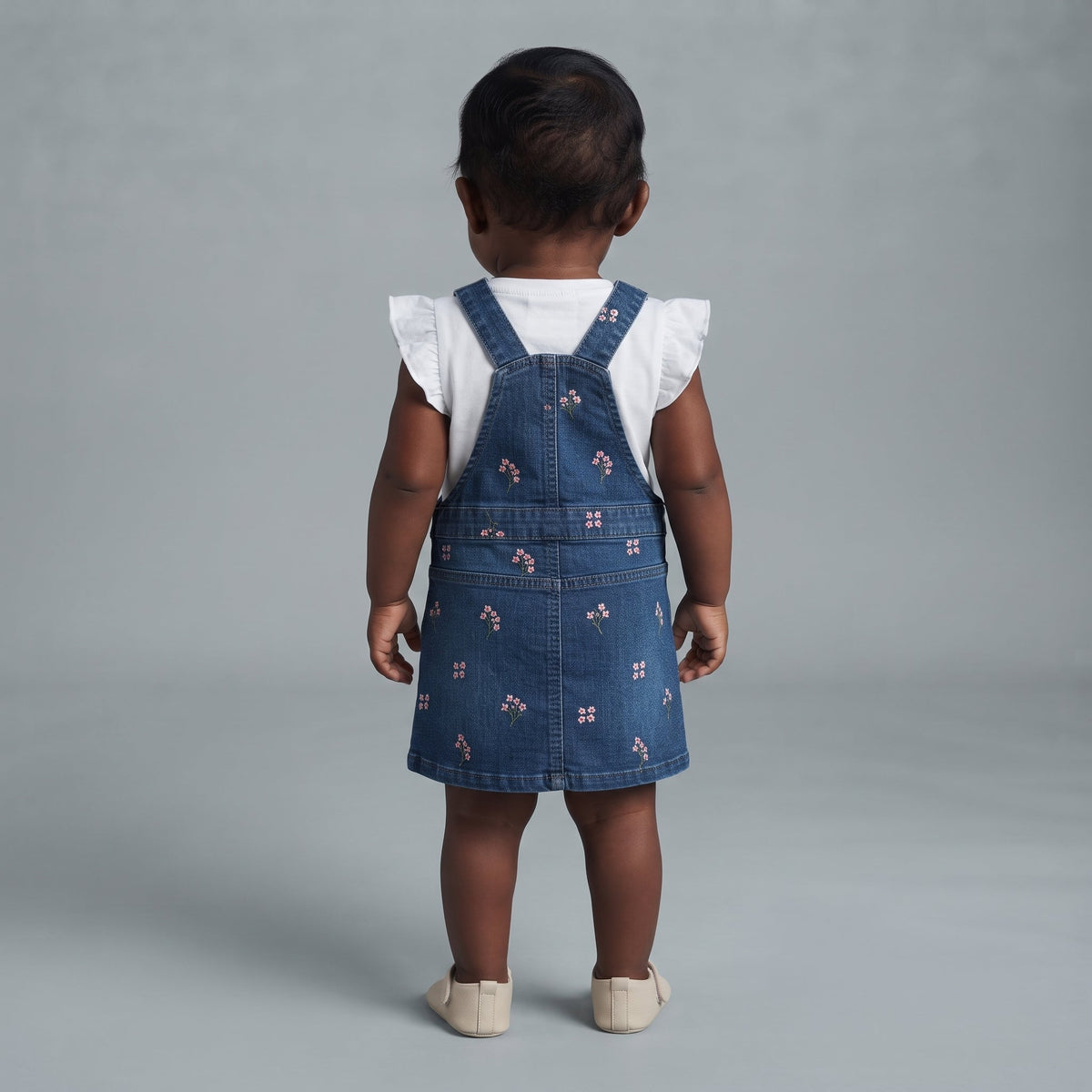 Bebé niña vistiendo un overol de mezclilla EmpiBy con bordados florales y camisa blanca con volados, vista trasera.