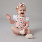 Bebé sonriente con conjunto EmpiBy de overol rosa estampado con flores, camiseta blanca con volantes y diadema con lazo