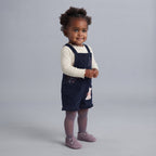 Niña pequeña con overol EmpiBy azul marino de pana, camiseta crema y zapatos lilas sobre fondo gris