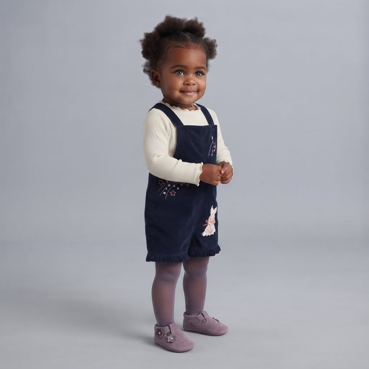 Niña pequeña con overol EmpiBy azul marino de pana, camiseta crema y zapatos lilas sobre fondo gris