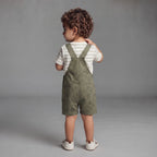 Niño pequeño de espaldas vistiendo overol corto verde oliva con estampado de Simba y camiseta a rayas de EmpiBy