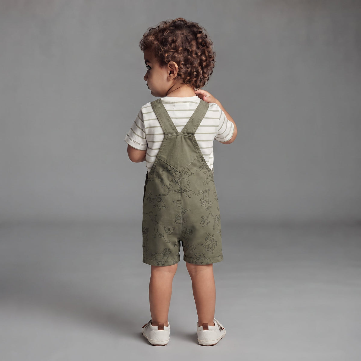 Niño pequeño de espaldas vistiendo overol corto verde oliva con estampado de Simba y camiseta a rayas de EmpiBy