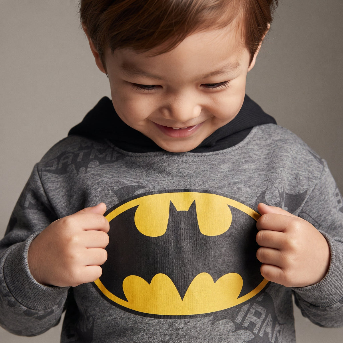 Niño sonriente vistiendo sudadera gris EmpiBy con capucha y logo de Batman en amarillo y negro