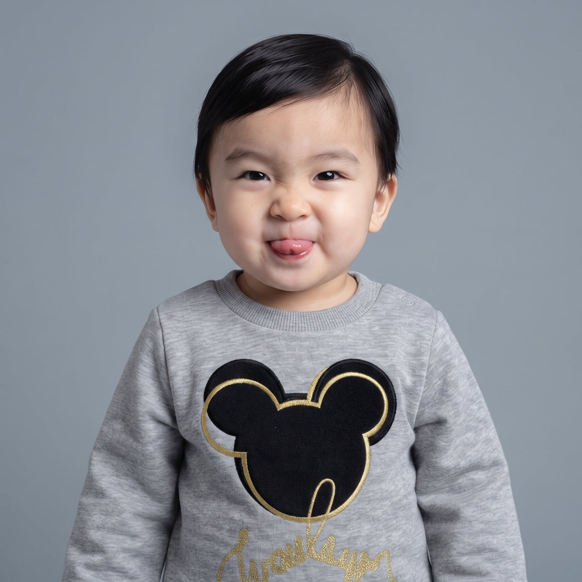 Bebé sonriente vistiendo sudadera gris EmpiBy con estampado de Mickey Mouse en negro y detalles dorados