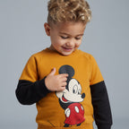 Niño sonriente vistiendo sudadera EmpiBy mostaza y negra con estampado de Mickey Mouse y mangas en contraste