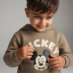 Niño sonriente vistiendo sudadera con capucha EmpiBy color topo con estampado de Mickey Mouse y letras crema