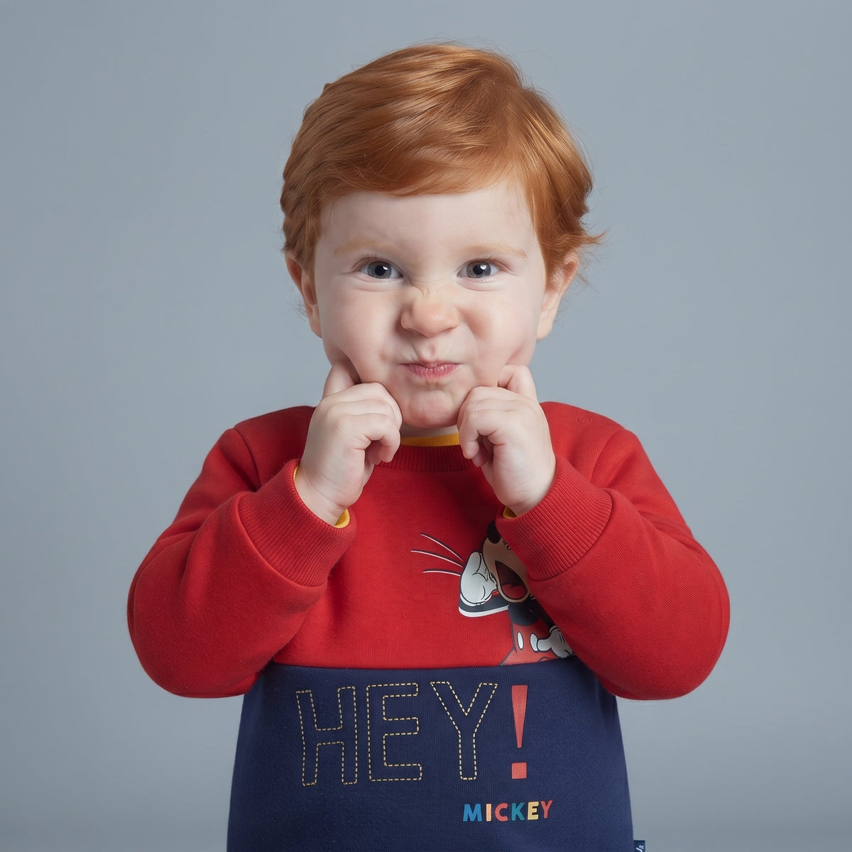 Niño pequeño con sudadera EmpiBy roja y azul de Mickey Mouse con estampado HEY! sobre fondo gris neutro