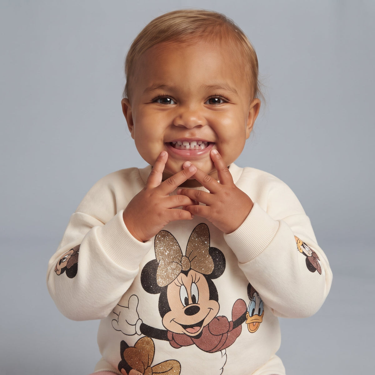 Bebé sonriente vistiendo sudadera EmpiBy color marfil con estampado brillante de Minnie Mouse y Daisy Duck
