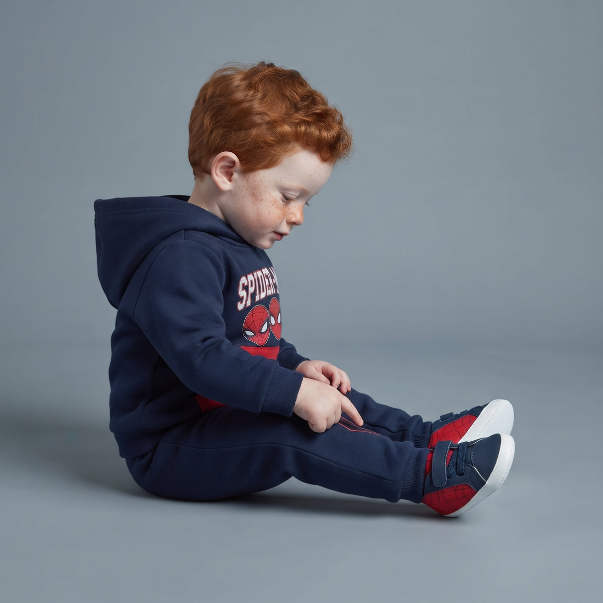 Bebé pelirrojo con sudadera azul de Spider-Man y pantalón jogger a juego de EmpiBy, sentado sobre fondo gris.