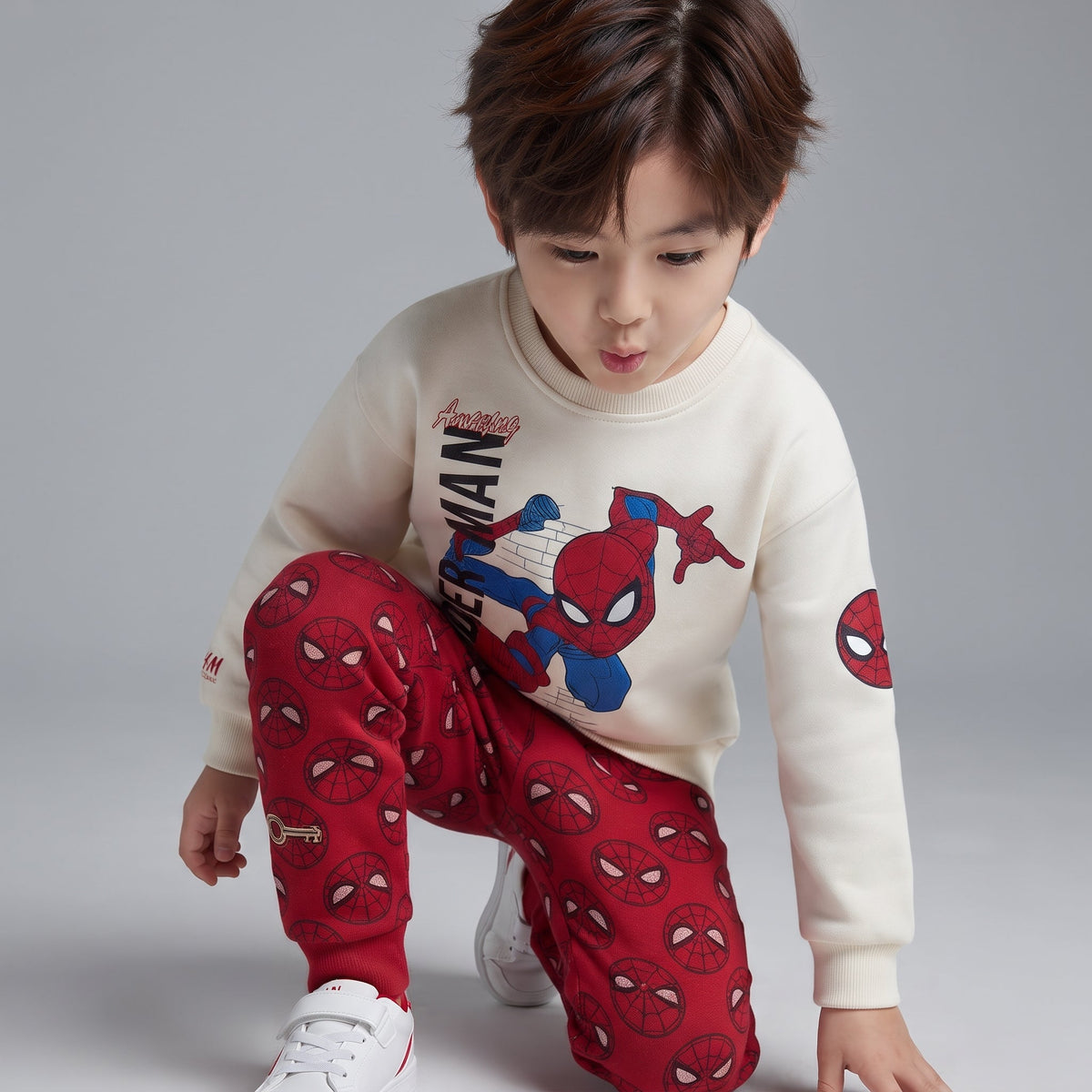 Niño con conjunto EmpiBy de dos piezas: sudadera beige de Spider-Man y pantalón jogger rojo con estampado de máscaras.