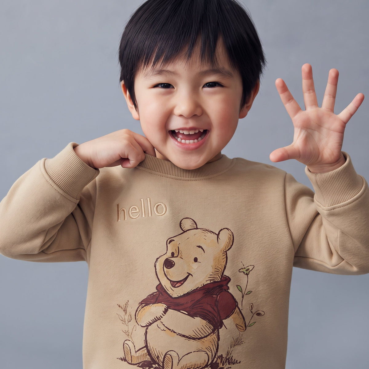 Niño sonriente vistiendo sudadera beige de EmpiBy con estampado de Winnie the Pooh y texto hello en el pecho