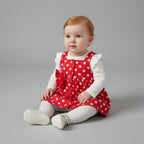 Bebé niña con conjunto EmpiBy de vestido peto rojo con lunares blancos, camiseta blanca y medias sobre fondo gris